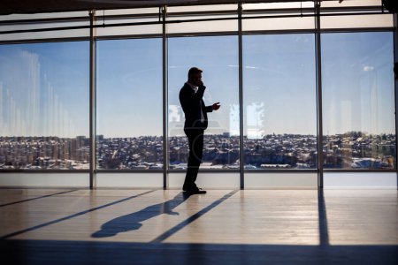 Rückansicht eines Geschäftsmannes, der aus einem großen Fenster auf die Stadt blickt. Er hält ein Telefon in der Hand. Horizontale Ansicht.