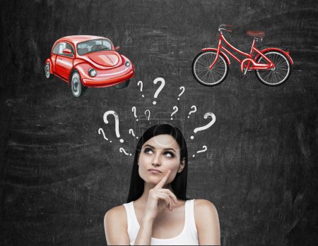 A beautiful brunette woman is trying to chose the most suitable way for travelling or commuting. Two sketches of a car and a bicycle are drawn on the black chalkboard.