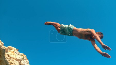 Téléchargez les photos : BOTTOM UP, CLOSE UP : Fit touriste mâle saute d'une haute falaise pour plonger dans l'océan rafraîchissant et se rafraîchir de la chaleur de l'été. Jeune homme caucasien en vacances d'été rock plongée par une journée ensoleillée. - en image libre de droit