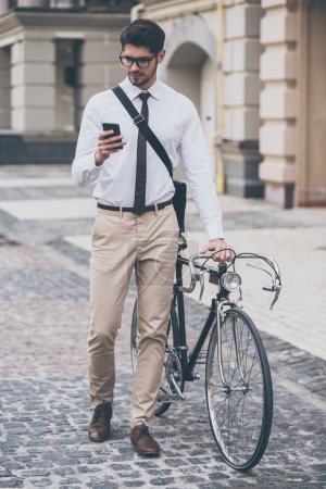 Téléchargez les photos : Message en route. Jeune homme confiant dans des lunettes utilisant son téléphone intelligent et tenant la main sur son vélo tout en se tenant debout à l'extérieur - en image libre de droit