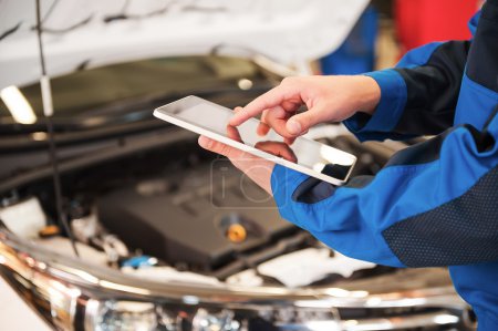 Téléchargez les photos : Gros plan de l'homme en uniforme travaillant sur tablette numérique tout en se tenant devant le capot de la voiture en atelier - en image libre de droit