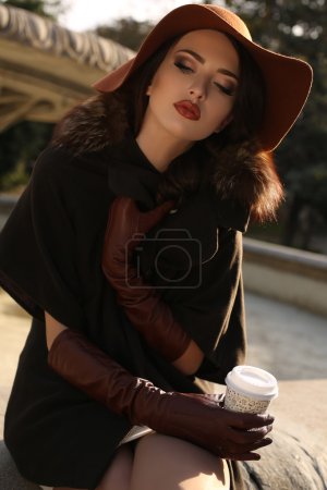 Foto de Foto de moda al aire libre de la hermosa dama con un abrigo elegante con piel, sombrero de fieltro y guantes de cuero, sosteniendo una taza de papel de café en la mano - Imagen libre de derechos