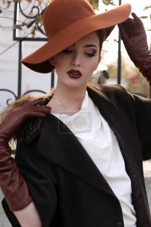 Foto de Foto de moda al aire libre de la hermosa dama con el pelo oscuro usando un abrigo elegante, guantes de cuero y sombrero de fieltro, posando en el parque de otoño - Imagen libre de derechos