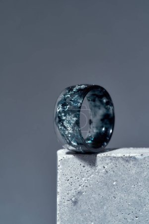 Primer plano del anillo hecho a mano de resina epoxi azul oscuro con lámina de plata en el interior sobre elemento de hormigón aislado sobre fondo gris Primer plano del anillo hecho a mano de resina epoxi azul oscuro con lámina de plata en el interior sobre elemento de hormigón aislado sobre fondo gris