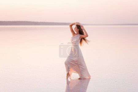 Young blonde woman in an evening airy pastel pink, powdery dress stands barefoot on white crystallized salt. Girl with natural make-up, hair is developing. Salt mining trip, walking on water at sunset