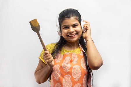 Eine hübsche indische Hausfrau mit Kochschürze und Kopfhörer mit Holzspachtel in der Hand lächelt auf weißem Hintergrund