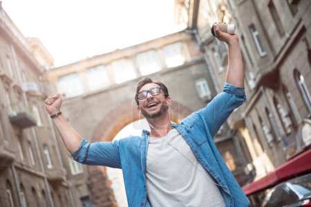 Photo for Absolute happiness. Joyful good looking young man walking down the street and holding his hands up while listening to music. - Royalty Free Image