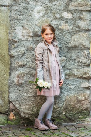 Außenporträt eines süßen kleinen Mädchens mit weißen Rosen in beigem Mantel und rosa Kleid