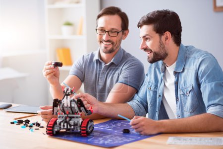 Foto de Trabaja con deleite. Compañeros sonrientes alegres sentados en la mesa y construyendo robot mientras trabajan en el proyecto - Imagen libre de derechos