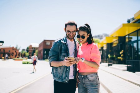 Millennial friends check online information about together web blog smiling during mobile networking, happy male and female Generation Z in trendy glasses watching media video via cellphone app