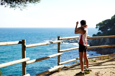 Téléchargez les photos : Plan latéral d'un beau jeune coureur étirant ses bras avant de commencer sa course en se tenant debout sur le bord d'une falaise avec une clôture en bois et profitant d'une vue sur l'océan depuis l'altitude - en image libre de droit