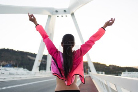 Téléchargez les photos : Vue arrière de la femme heureuse avec une silhouette élancée debout sur un pont de la ville avec les mains levées tout en profitant d'une belle journée après l'entraînement, jeune femme sportive vêtue de survêtement se détendre après l'entraînement - en image libre de droit