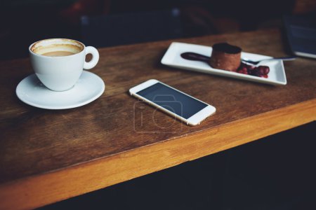 Téléchargez les photos : Gros plan de téléphone portable couché sur une table en bois près de la tasse de cappuccino et assiette de dessert sucré à l'intérieur du café, concept de temps libre, repos dans le café - en image libre de droit