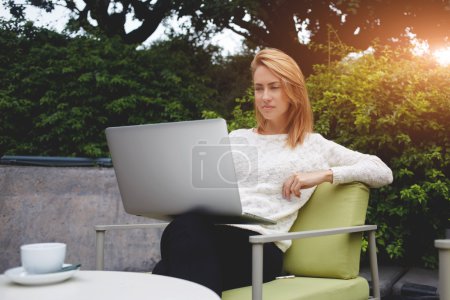 Photo for Young charming woman watching video on laptop computer while relaxing in sidewalk cafe during coffee break, beautiful female reading fashion news on net-book while sitting in restaurant outdoors - Royalty Free Image