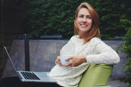 Foto de Jovencita alegre sosteniendo taza de capuchino mientras está sentada con netbook portátil en la acogedora cafetería de la acera, chica hipster bastante sonriente disfrutando del café mientras descansa después del trabajo en su computadora portátil - Imagen libre de derechos