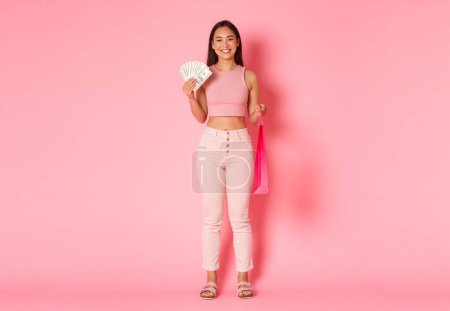 Foto de Longitud completa de atractiva chica asiática sonriente en ropa de verano, sosteniendo el dinero y la bolsa de compras, la compra de algo caro, disfrutando de caminar en los centros comerciales, de pie fondo rosa. - Imagen libre de derechos
