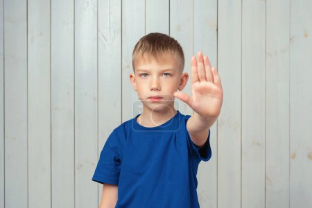 Yo digo que no. Niño preadolescente serio en camiseta azul mantiene la palma hacia adelante en la cámara, restringe o advierte, extiende el brazo en señal de stop. Estudio plano, fondo de madera claro Yo digo que no. Niño preadolescente serio en camiseta azul mantiene la palma hacia adelante en la cámara, restringe o advierte, extiende el brazo en señal de stop. Estudio plano, fondo de madera claro