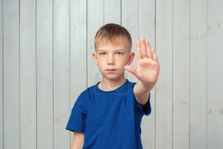 I say no. Serious preteen boy in blue t shirt keeps palm forward at camera, restricts or warns, extends arm in stop sign. Studio shot, light wooden background