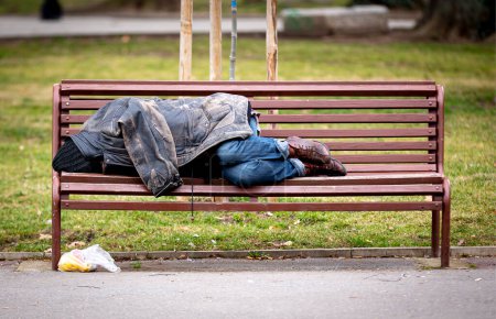 Hombre sin hogar duerme en un banco cubierto con su chaqueta en un parque público durante el día.