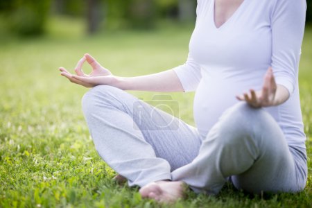 Foto de Primer plano del torso de la joven modelo embarazada trabajando en el parque. Futura mamá sentado piernas cruzadas en fácil postura de yoga en la sesión de meditación en césped de hierba. Yoga prenatal. Concepto de maternidad saludable - Imagen libre de derechos