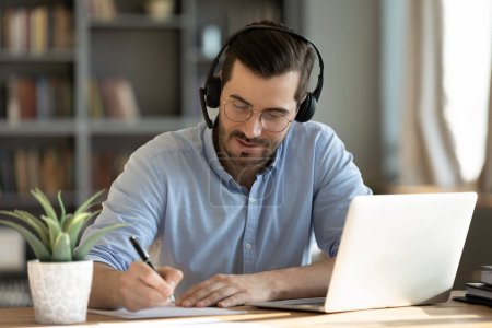 Foto de El hombre enfocado de 35 años se sienta en el escritorio, usa auriculares, mira webinar, usa computadoras portátiles, gana nuevos conocimientos escribiendo notas. Comunicación por videoconferencia, negociación remota, estudio en línea, e-learn, autoeducación concepto - Imagen libre de derechos