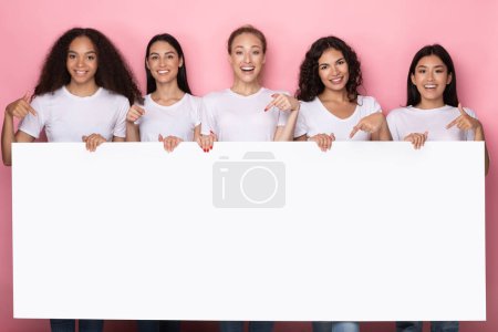 Mujeres sosteniendo tablero de papel vacío de pie sobre fondo de estudio rosa