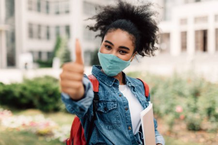 Schwarzes Studentenmädchen mit Gesichtsmaske gestikuliert Daumen hoch im Freien