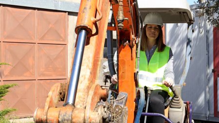 Téléchargez les photos : Une femme contremaître portant un casque de travail et une veste réfléchissante conduit une pelle sur le chantier. Concept de travail. - en image libre de droit