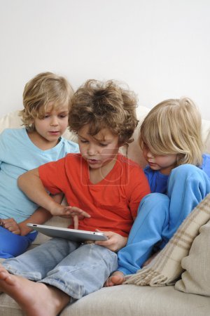 Téléchargez les photos : Un enfant de 8-9 ans joue à un jeu sur une tablette tactile. Ses frères cadets surveillent chaque mouvement intensément - en image libre de droit