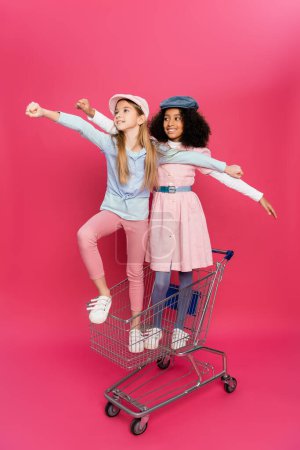 fashionable, happy interracial girls posing in shopping cart on pink