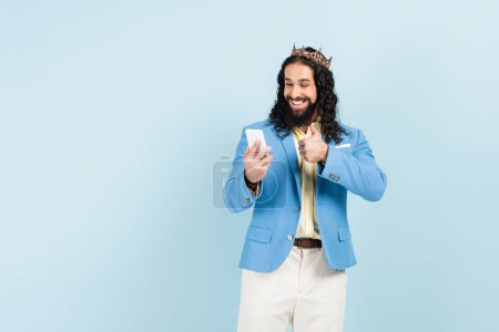 bearded hispanic man in jacket and crown showing thumb up during video call isolated on blue