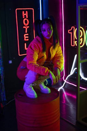 Photo for Brunette asian woman sitting near neon sign with hotel lettering outside - Royalty Free Image