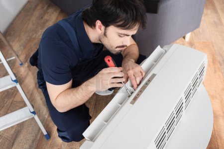 high angle view of bearded handyman holding screwdriver while fixing broken air conditioner