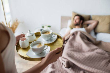 Frau hält Tablett mit Tassen Kaffee in der Nähe verschwommener und schlafender Mann 