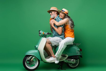 cheerful and interracial middle aged couple in sun hats hugging while riding moped on green