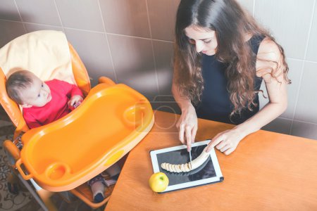 Foto de Madre cocinando alimentos para bebés en la computadora de la tableta PC. Mamá confundida erróneamente con la tabla de cortar. Cristal fuerte, irrompible, pantalla a prueba de roturas. Protector. Impermeable. Receta. Internet. - Imagen libre de derechos