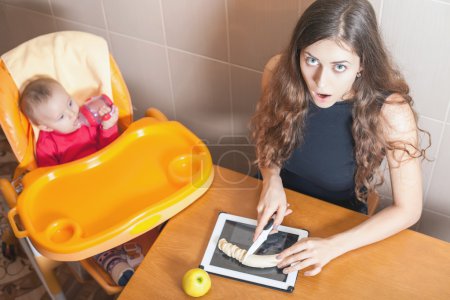 Foto de Madre cocinando alimentos para bebés en la computadora de la tableta PC. Mamá confundida erróneamente con la tabla de cortar. Cristal fuerte, irrompible, pantalla a prueba de roturas. Protector. Impermeable. Receta. Internet. - Imagen libre de derechos