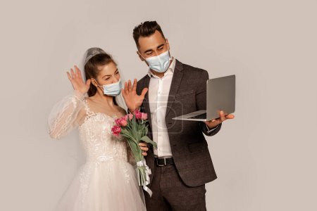 bride and groom in medical masks waving hands during video call isolated on grey bride and groom in medical masks waving hands during video call isolated on grey