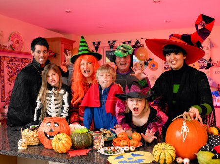 Téléchargez les photos : Famille / amis posant pour la caméra dans leurs costumes d'Halloween. Ils sont debout dans la cuisine avec de la nourriture de fête et des friandises exposées devant eux
. - en image libre de droit