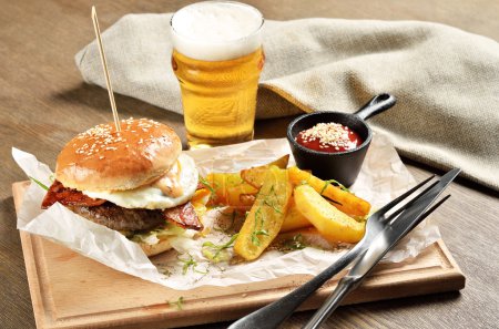 Photo for Burger, mexican potatoes and ketchup on wooden board. Glass of beer in the background. - Royalty Free Image