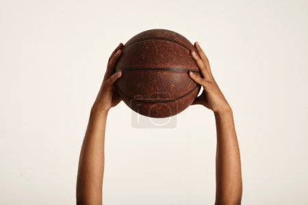 Photo for Close up shot of two strong dark-skinned hands holding up an old vintage leather brown basketball, isolated on white - Royalty Free Image