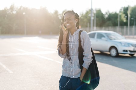 Photo for Beautiful african american woman walkin with hand in pocket talking on smart phone in city, looking away, outdoors sunny exterior. - Royalty Free Image