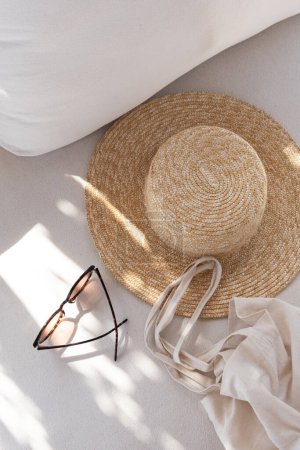 Pisos de accesorios de moda para mujer. Elegantes gafas de sol femeninas, sombrero de paja, bolso de compras en un sofá salón blanco con almohadas. Vista superior