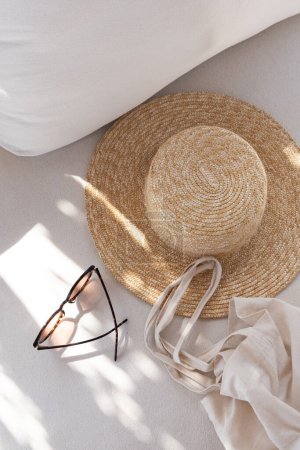 Téléchargez les photos : Flatlay d'accessoires de mode pour femmes. Lunettes de soleil féminines élégantes, chapeau de paille, sac à provisions sur canapé salon blanc avec oreillers. Vue du dessus - en image libre de droit