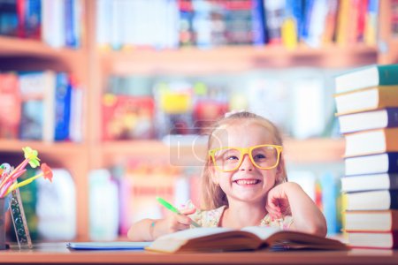 Photo for Clever little schoolgirl in glasses at table with books trying to learn new skills - Royalty Free Image