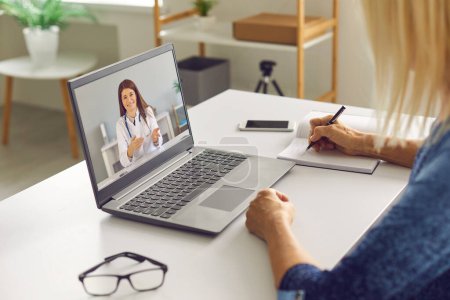 Reife Frau macht sich Notizen, während sie einem Online-Arzt zuhört, der Gesundheitsratschläge gibt