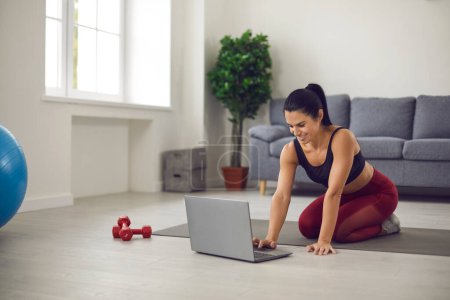 Frau sitzt zu Hause auf Sportmatte, kauft online Sportbekleidung ein oder schaut sich Video-Trainingsstunde an